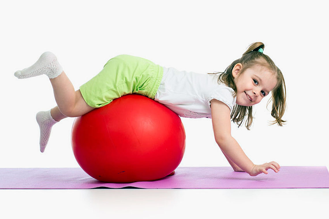 Criança a baloiçar sobre grande bola vermelha, em aula de fisioterapia infantil.